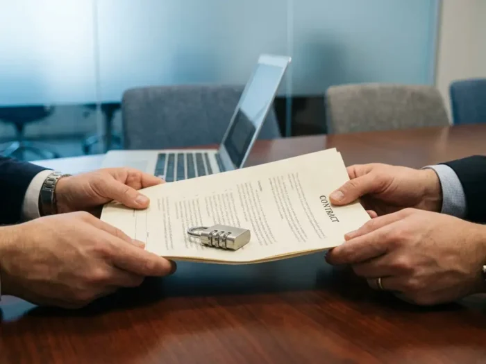 Twee handen houden een formeel contract vast op een zakelijk bureau, met een cijferslot bovenop de documenten en een laptop op de achtergrond.