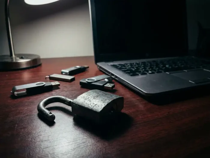 Gebroken stalen hangslot op donker bureau naast USB-sticks en laptop, symboliseert cyberbeveiliging en datalek.