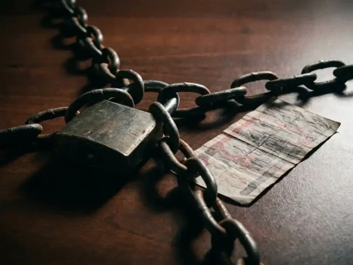 Verroest hangslot omwikkeld met zware kettingen naast verkreukeld bankbiljet op donker bureau, dramatische zijverlichting.