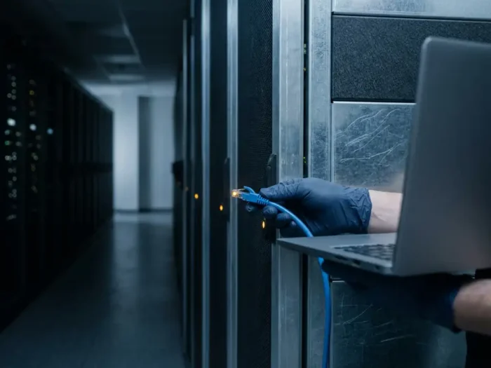 Gehandschoende hand verbindt laptop met serverrack via ethernetkabel in donkere datacentergang met blauwstalen tonen.